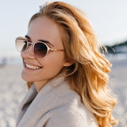 Close-up portrait of positive girl with wavy blonde hair dressed in beige cashmere sweater and sunglasses on beach.