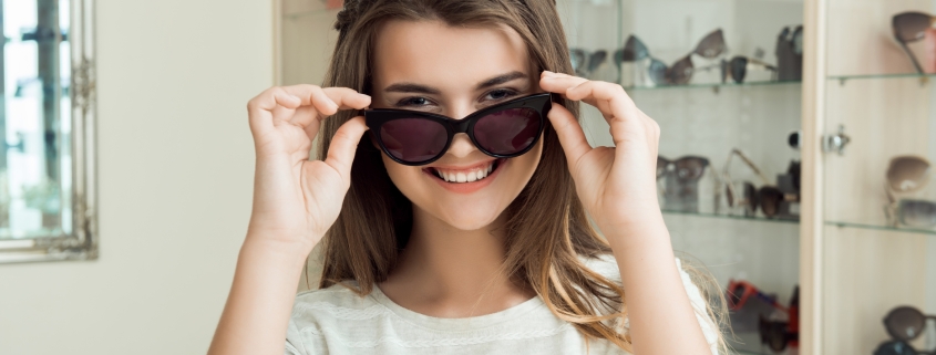 Pretty brunette girl smiling, trying on sunglasses in optical shop, asking boyfriend if they suit her, trying to find perfect eyewear before going on vacation to sea. Girl in store enjoys shopping